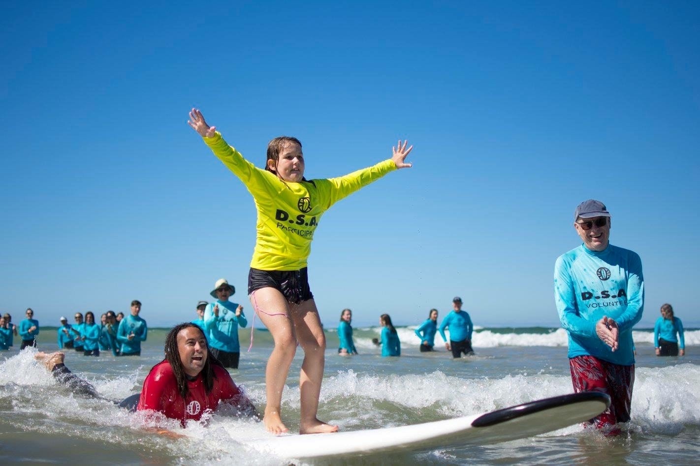A participant standing triumphantly on a surfboard with arms raised — supported by DSA volunteers